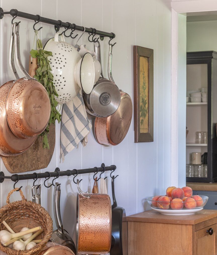 Copper and stainless steel cookware hanging from black metal rails on a white paneled wall.