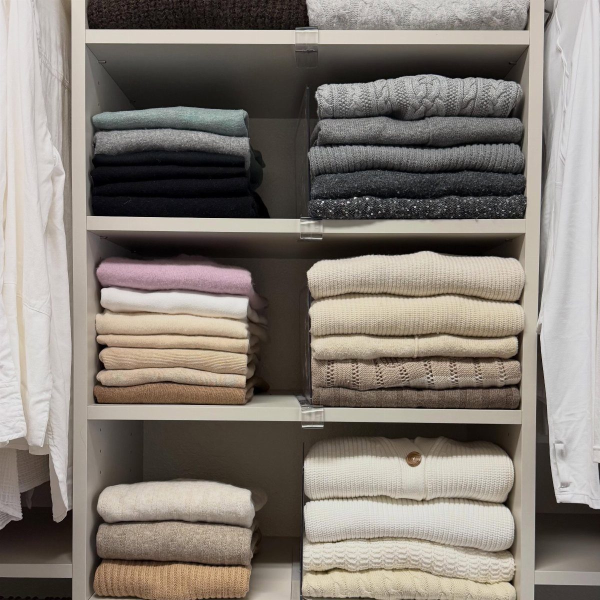 Folded sweaters in neutral colors stacked neatly on white open closet shelves.