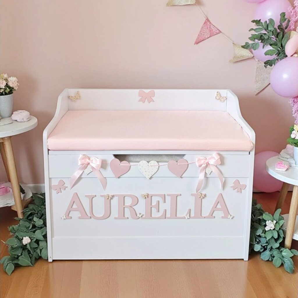 White wooden toy chest with pink name lettering and padded lid in a nursery.
