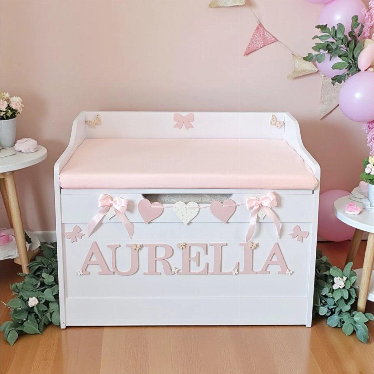 White wooden toy chest with pink name lettering and padded lid in a nursery.