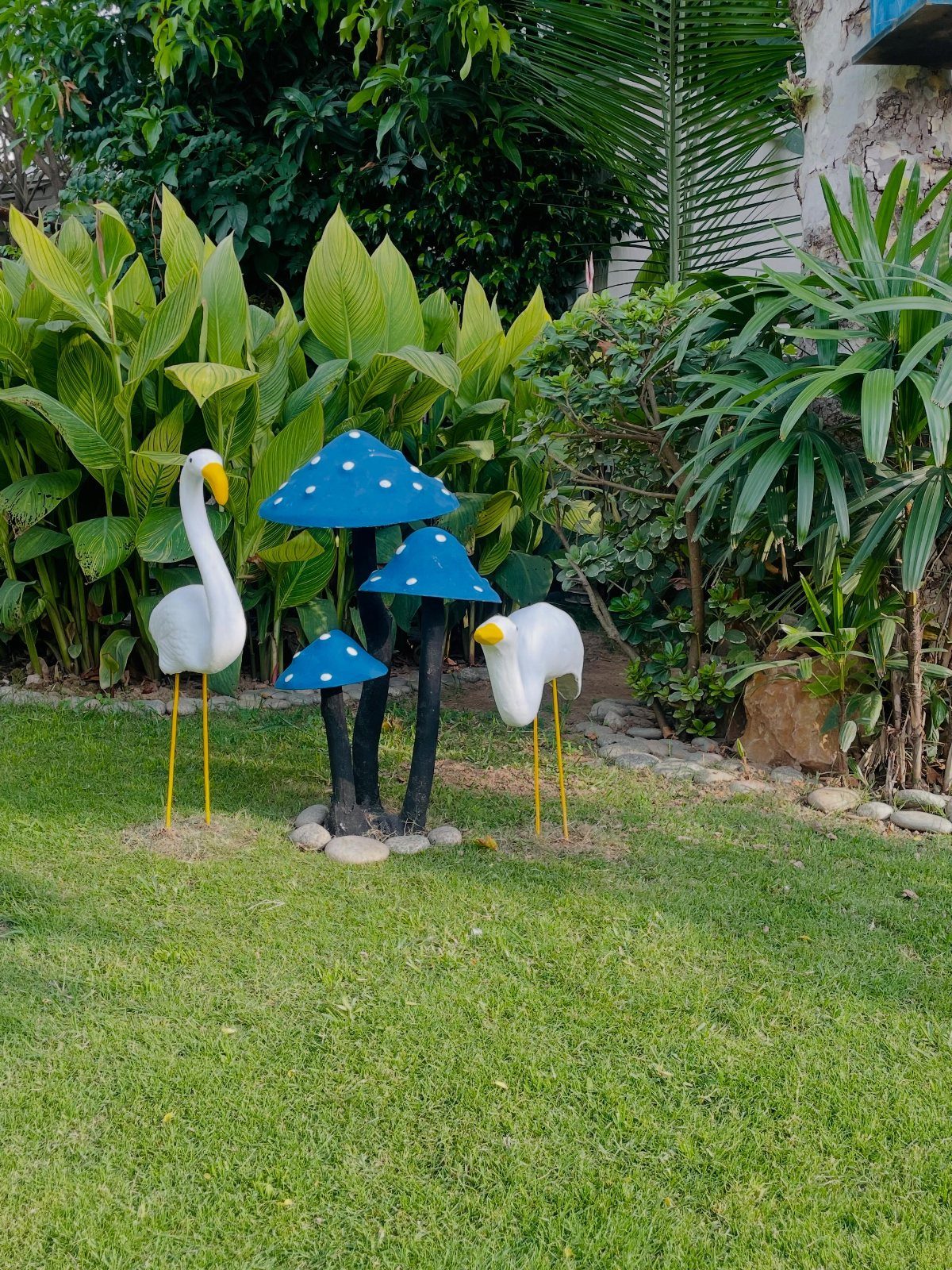 Blue mushroom sculptures and white bird statues standing on a green lawn surrounded by tropical plants.