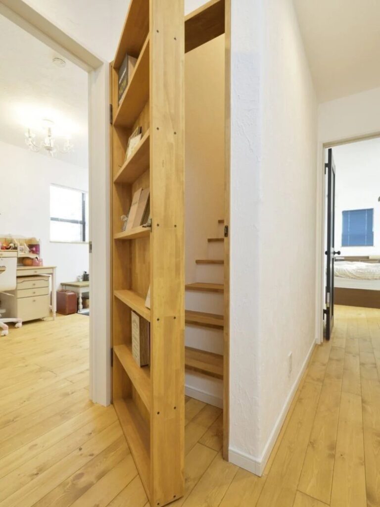 A wooden bookcase door swings open to reveal a hidden staircase in a bright hallway.