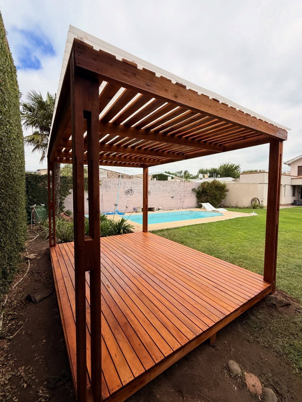 A wooden deck with a slatted pergola overlooking a swimming pool and green lawn.
