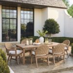 Wooden dining table with woven chairs and white cushions on a stone patio.