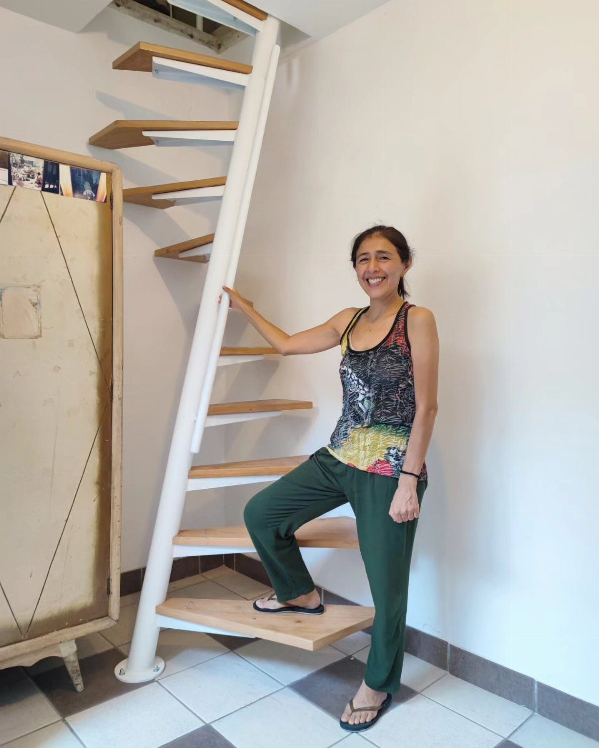 A smiling woman stands beside a white spiral staircase with wooden steps in a bright room.
